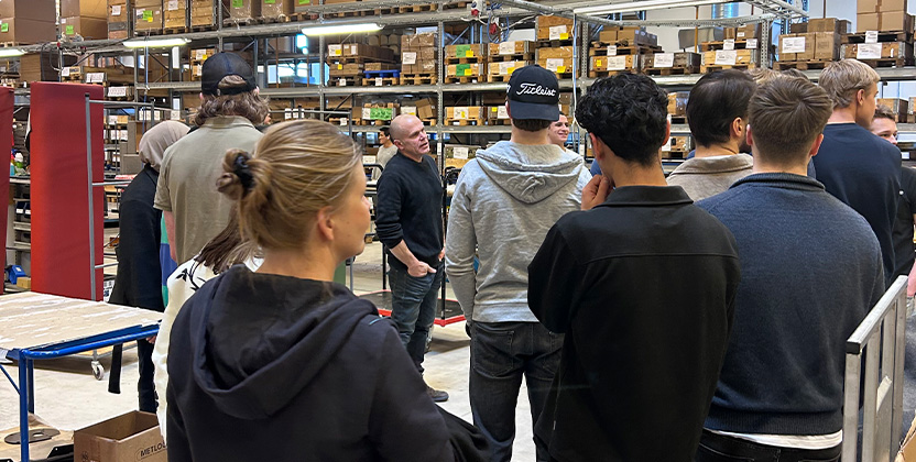 En grupp elever på studiebesök i lagermiljö. En person guidar och berättar.