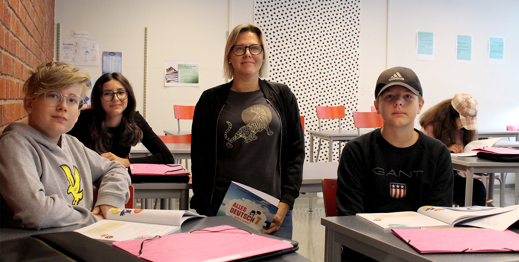 I ett klassrum sitter tre elever, två pojkar och en flicka, vid sina skolbänkar och deras kvinnliga lärare står mellan deras bänkar och håller i en tysk kursbok. De tittar in i kameran. 