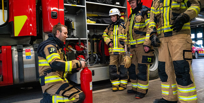Fyra brandmän i full utrustning står vid en brandbil och hanterar brandsläckare i en brandstation.