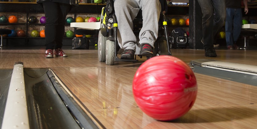 Ett rött bowlingklot far mot kameran. I bakgrunden finns tre personer, varav en sitter i rullstol.