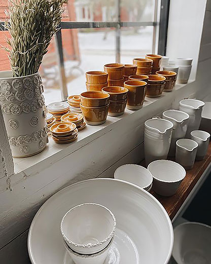 Vita och bruna skålar, krukor, karaffer och ljusstakar i keramik står uppställda i och framför ett fönster.