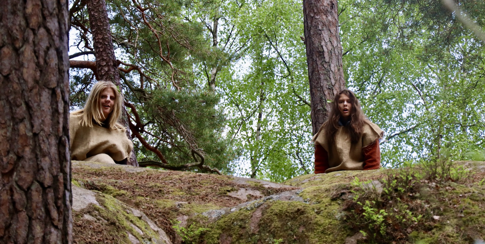 Två tjejer i 18-årsåldern utklädda till rumpnissar gömmer sig bakom träd och stenar i skogen. 
