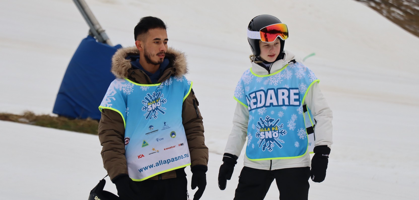 En tjej och en kille i 18-årsåldern står i en skidbacke klädda i skidkläder, utrustning samt västar med texten "Alla på snö"