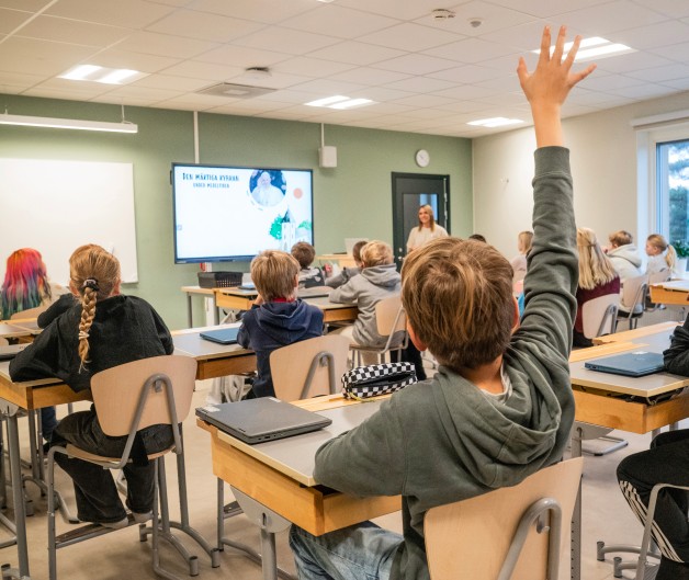 Ett klassrum med elever som sitter vid sina bänkar, en elev räcker upp handen och en lärare står framme vid tavlan.