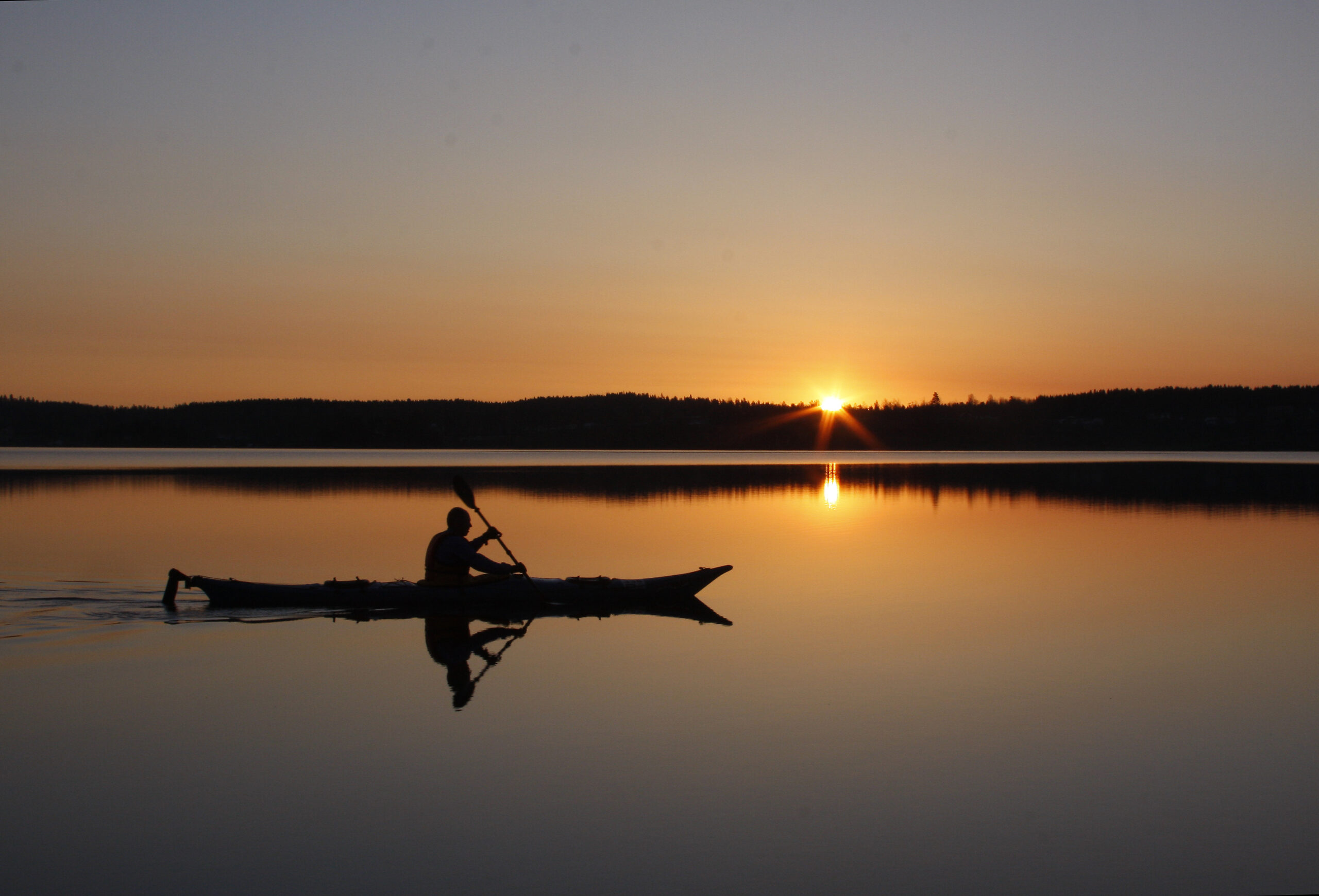 En person paddlar kajak på en stilla sjö vid solnedgången, med spegling i vattnet.
