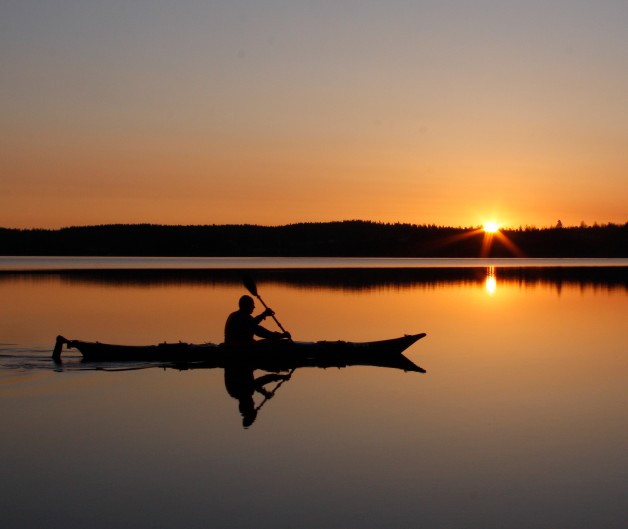En person paddlar kajak på en stilla sjö vid solnedgången, med spegling i vattnet.