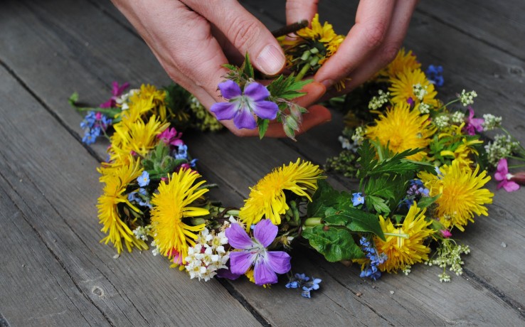 Händer flätar en blomkrans av gula, lila och vita blommor på ett trägolv.