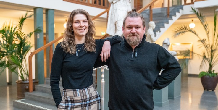 Två personer står bredvid varandra i en ljus, modern foajé med trappor och statyer i bakgrunden.