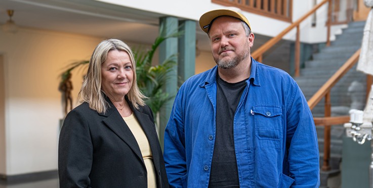 Två personer står tillsammans inomhus, nära en trappa och växter. Ansikten är suddade.