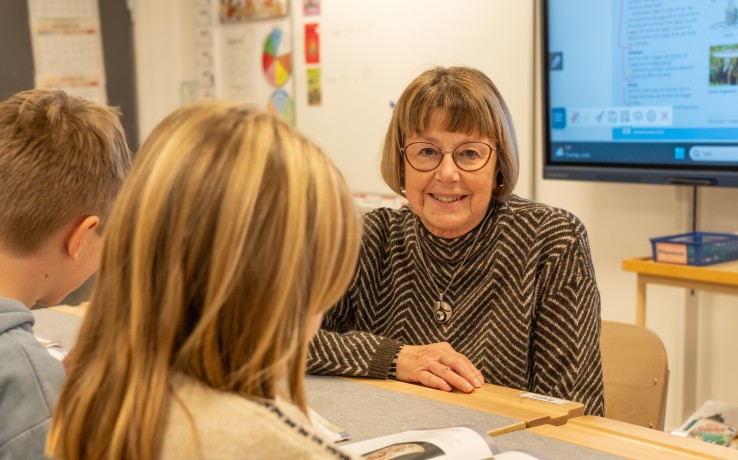 Två barn och en vuxen sitter vid ett bord i ett klassrum och arbetar tillsammans med en bok.