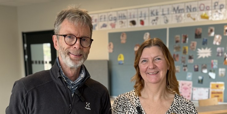 Två vuxna står i ett klassrum med ansiktena suddade, omgivna av skolbänkar och en anslagstavla.