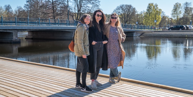 Tre personer står på en brygga vid vattnet med en bro och träd i bakgrunden, soligt väder.