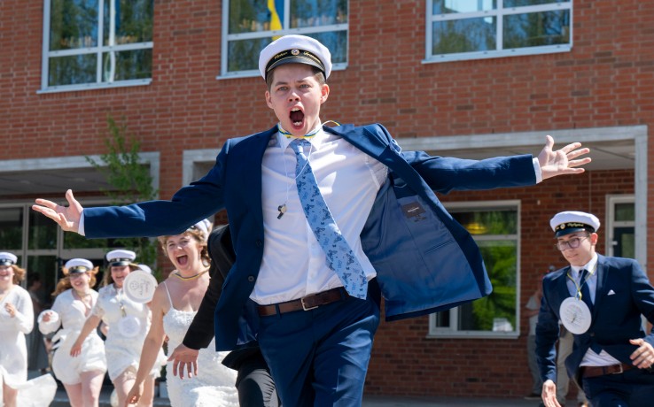 Studenter i vita mössor och festkläder springer glada ut från en skolbyggnad.