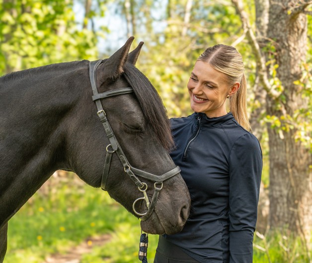 Kvinna i svart tröja står bredvid en svart häst i en grön skogsmiljö.