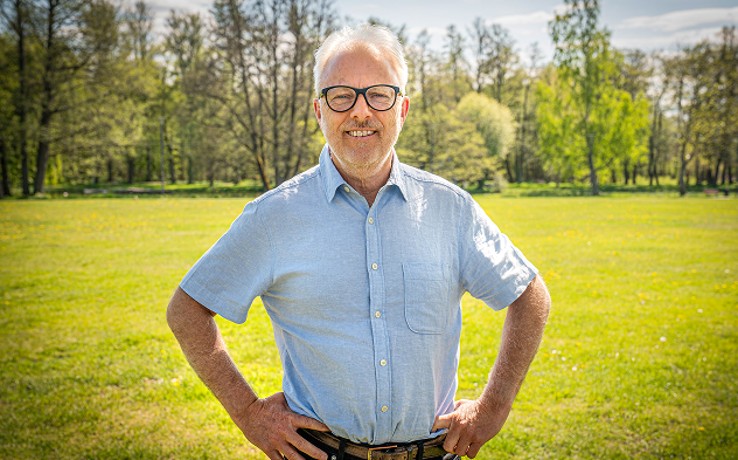En man med grått hår står utomhus på en gräsmatta med händerna i sidan, ansiktet är suddat.