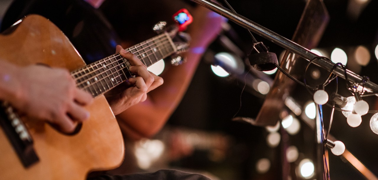 Närbild på en person som spelar akustisk gitarr på en scen med ljusslingor i bakgrunden.