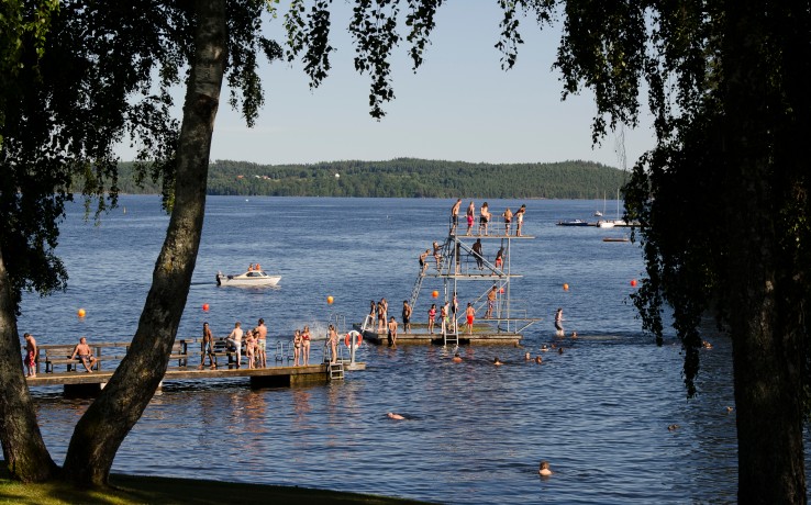 Människor badar, simmar och hoppar från hopptorn vid en sjö en solig sommardag.