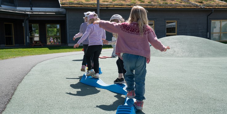 Flera barn balanserar på en blå lekbana utomhus vid en förskola en solig dag.