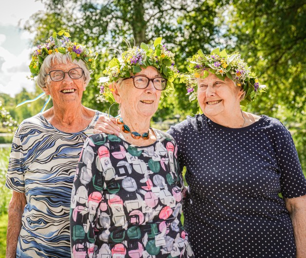 Tre äldre kvinnor med blomsterkransar står tillsammans utomhus i en grönskande park.