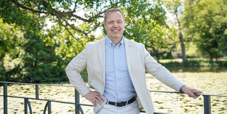 Man i ljus kostym står vid ett räcke vid en sjö, omgiven av grönska en solig dag.