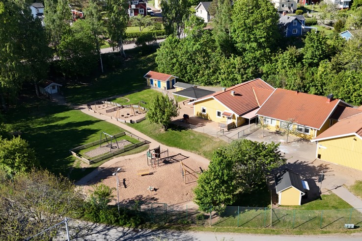 Fågelperspektiv av en förskola med lekplats, omgiven av grönska och bostadshus.