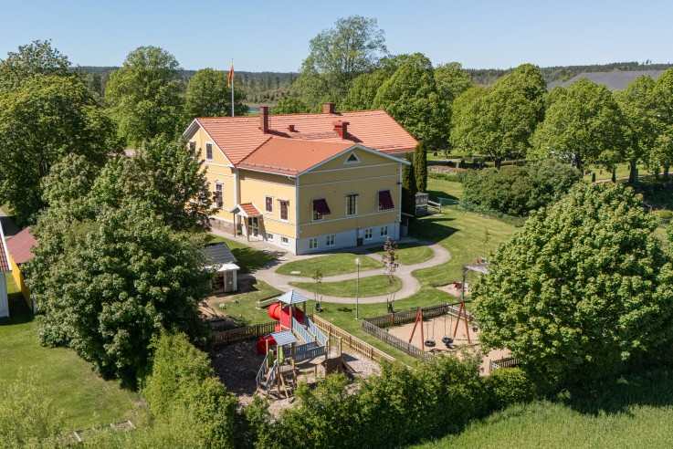 Fågelperspektiv över en gul byggnad med lekplats, omgiven av träd och gräsmattor en solig dag.