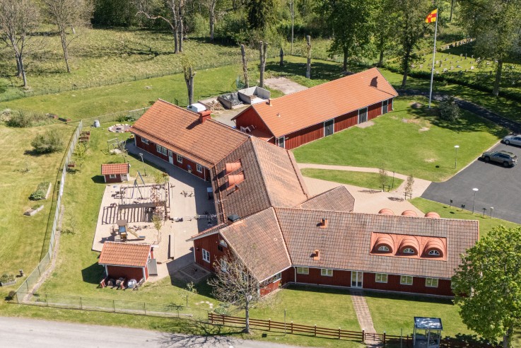 Flygbild över en lantlig förskola med röda byggnader, lekplats och omgivande grönska.