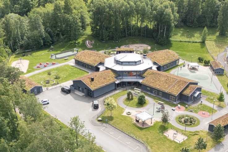 Flygbild över en modern skolbyggnad med gröna tak, omgiven av lekplatser, träd och grönområden.