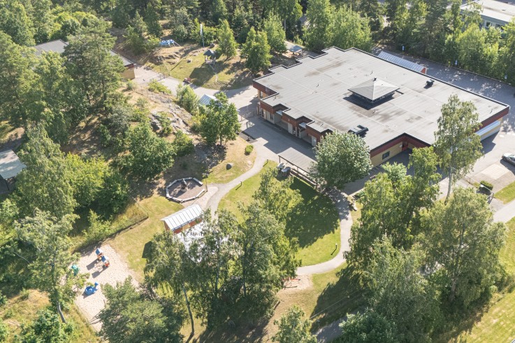 Flygbild över en byggnad omgiven av grönska, gångvägar och närliggande bostadshus.