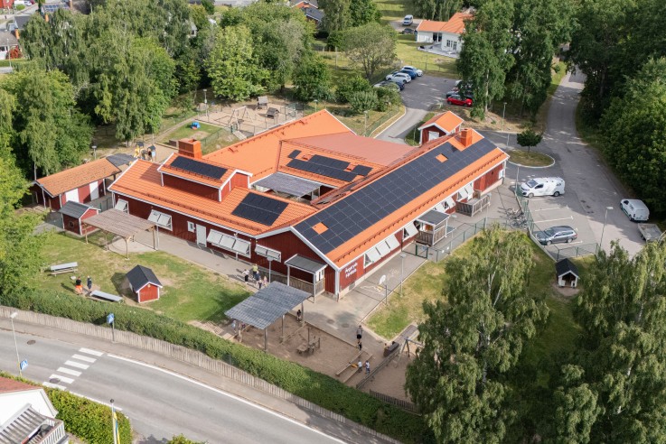 Flygbild över ett bostadsområde med röda hus, skola med solpaneler och grönska.