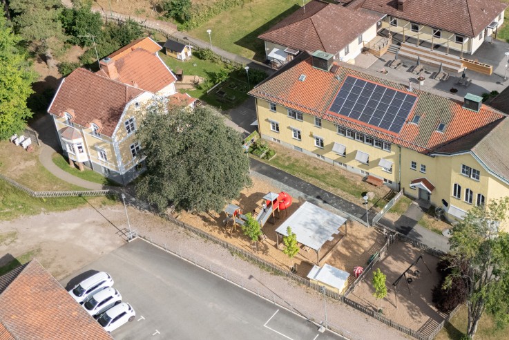 Flygbild över en skolbyggnad med solpaneler, lekplats, parkering och omgivande grönska.
