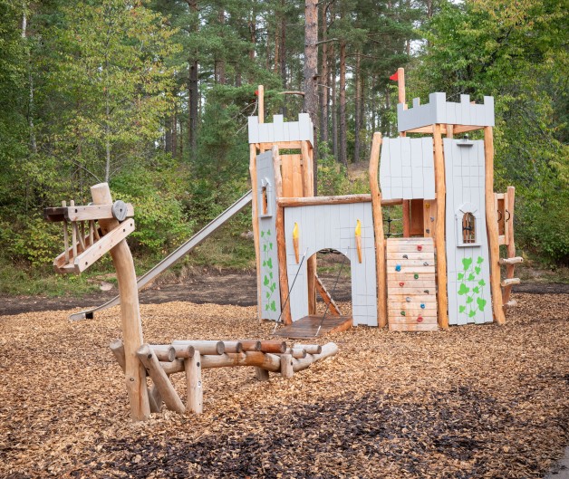 Lekplats med en borg i trä och en drakgunga, omgiven av skog och träflis på marken.