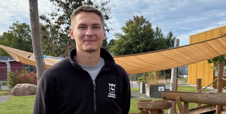 En man står utomhus på en lekplats med sand och träkonstruktioner, klädd i svart jacka.