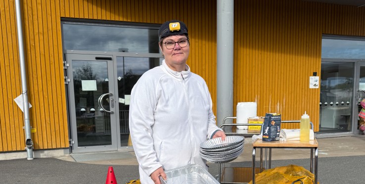 En person i vit jacka och svart mössa står utomhus vid en grillplats med aluminiumformar och en vagn med matvaror.