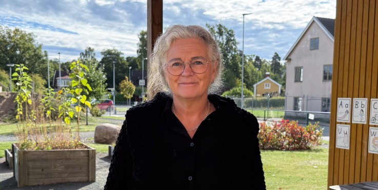 Kvinna med glasögon och grått hår står utomhus vid ett träbord, med hus och grönska i bakgrunden.
