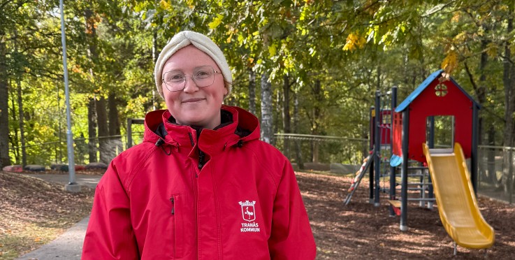 Person i röd jacka står utomhus nära lekplats och träd, höstlöv på marken.
