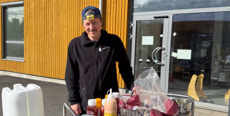 Man står utomhus vid en serveringsvagn med bröd, såser och andra grilltillbehör framför en gul träbyggnad.