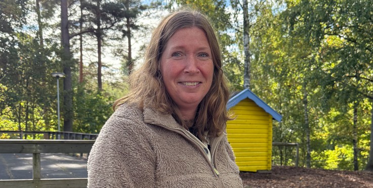 Kvinna i beige fleecejacka står utomhus framför ett gult skjul och träd en solig dag.