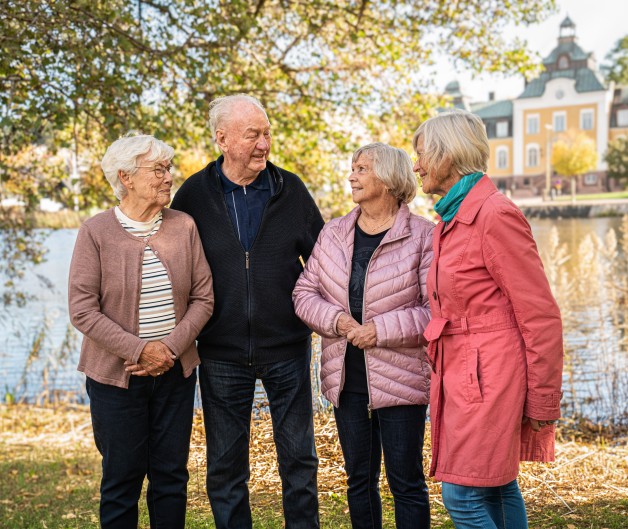 Fyra äldre personer står och samtalar utomhus vid en sjö, med träd och byggnad i bakgrunden.