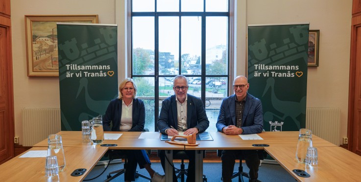 Tre personer sitter vid ett konferensbord framför fönster och två gröna banners med texten 'Tillsammans är vi Tranås'.
