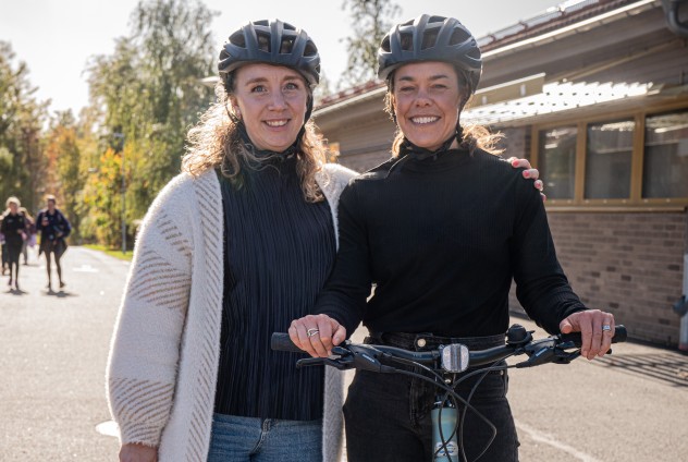 Två kvinnor med cykelhjälmar står utomhus, en håller i en cykel. Soligt väder, fler personer i bakgrunden.