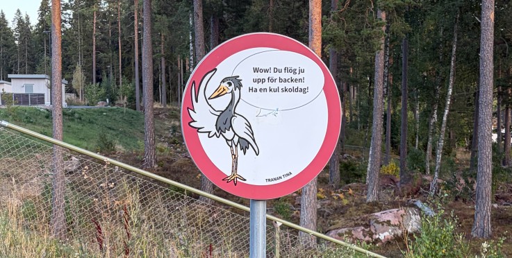 Rund skylt med tecknad trana och text på svenska, placerad vid en skogsstig nära ett stängsel.
