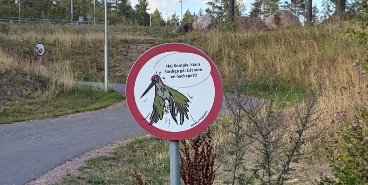 Vägskylt med tecknad hackspett och svensk text på en gångväg i naturmiljö med träd och gräs.