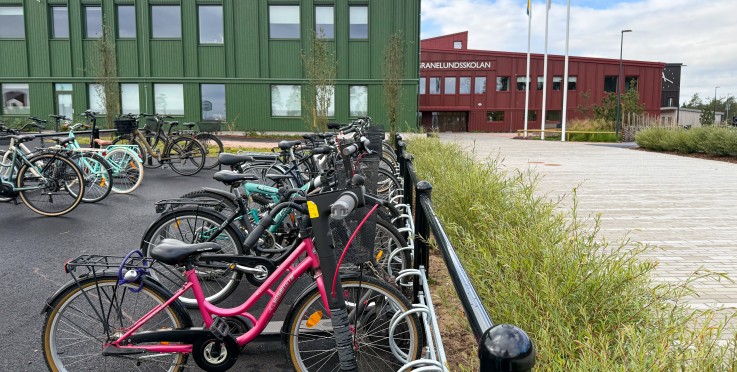 Cykelställ med flera cyklar utanför en grön och röd skolbyggnad, flaggstänger och gångväg syns.