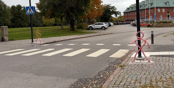 Övergångsställe med två röda figurer av metall, vägskyltar och en tegelbyggnad i bakgrunden.