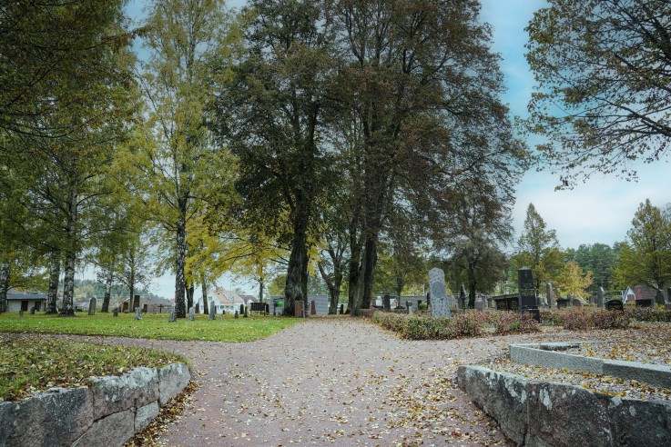 En lugn kyrkogård med gravstenar, träd och gångväg täckt av höstlöv under en mulen himmel.