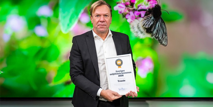 Man i kostym håller ett diplom framför en färgglad bakgrund med en blomma och en humla.