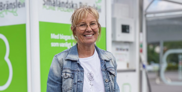 Leende kvinna i röda byxor och jeansjacka står framför en biogasmack utomhus.