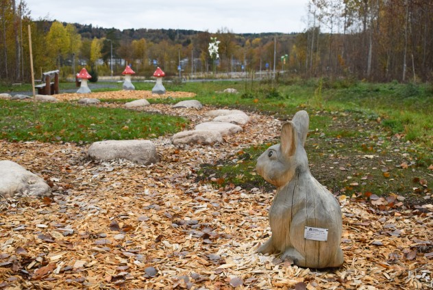 En träskulptur av en kanin står på en stig täckt av träflis i en park med höstfärger.